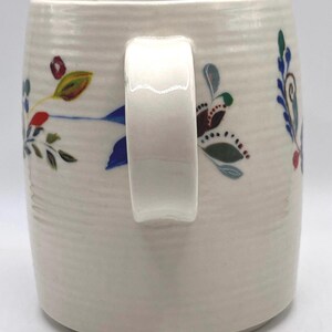 May include: Cream ceramic mug with a white handle and floral patterns. The mug features hand-painted flowers in blue, red, green, and brown. The mug has a textured, ribbed surface.