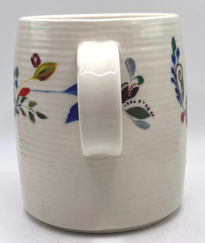 May include: Cream ceramic mug with a white handle and floral patterns. The mug features hand-painted flowers in blue, red, green, and brown. The mug has a textured, ribbed surface.