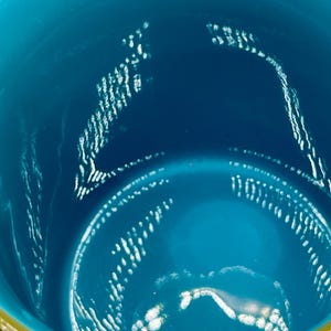 May include: Close-up of a teal-colored vessel with a gold rim. The interior of the vessel has a glossy finish with white, textured patterns. The gold rim adds a contrasting accent to the teal color.