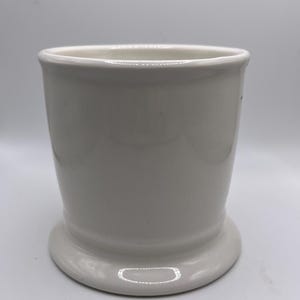 Puede incluir: Un vaso de cer&aacute;mica blanco con una base redondeada y una parte superior ligeramente ensanchada. El vaso tiene un acabado liso y brillante y un dise&ntilde;o simple y cl&aacute;sico. Es una pieza vers&aacute;til adecuada para diversos usos.