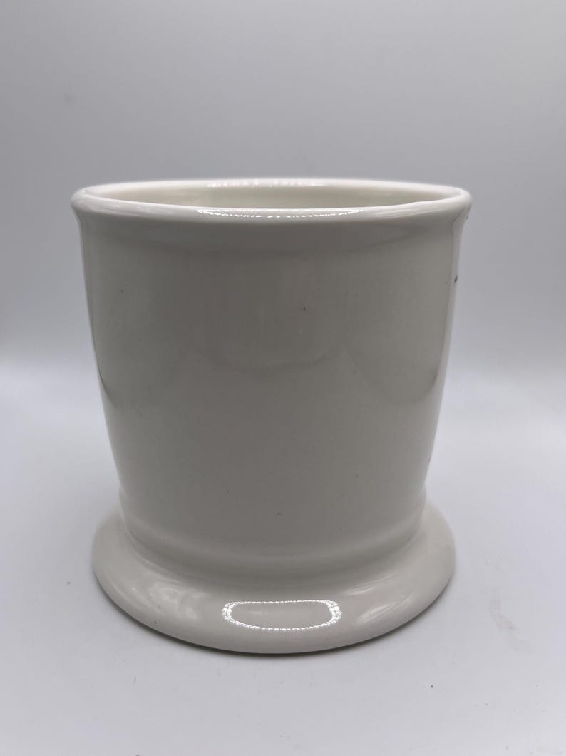 Puede incluir: Un vaso de cer&aacute;mica blanco con una base redondeada y una parte superior ligeramente ensanchada. El vaso tiene un acabado liso y brillante y un dise&ntilde;o simple y cl&aacute;sico. Es una pieza vers&aacute;til adecuada para diversos usos.