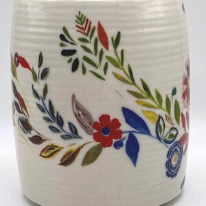 May include: A white ceramic mug with a floral design. The mug features a hand-painted pattern of colourful flowers and leaves in red, blue, green, and yellow. The mug has a cylindrical shape and a textured surface.