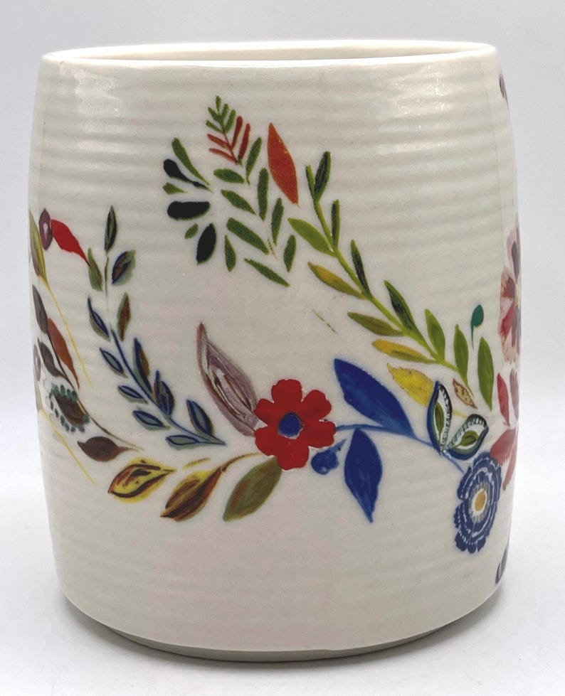 May include: A white ceramic mug with a floral design. The mug features a hand-painted pattern of colourful flowers and leaves in red, blue, green, and yellow. The mug has a cylindrical shape and a textured surface.