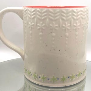 May include: Cream-colored ceramic mug with a red interior and a curved handle. The mug features embossed leaf and star patterns, with a green and blue floral design at the base. The mug is speckled with dark flecks.