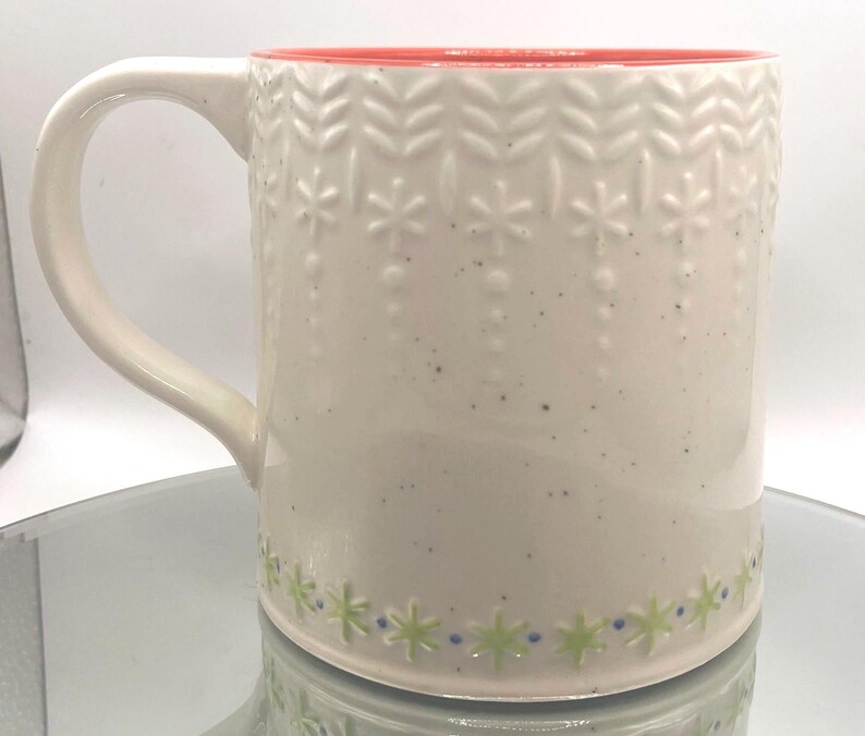 May include: Cream-colored ceramic mug with a red interior and a curved handle. The mug features embossed leaf and star patterns, with a green and blue floral design at the base. The mug is speckled with dark flecks.