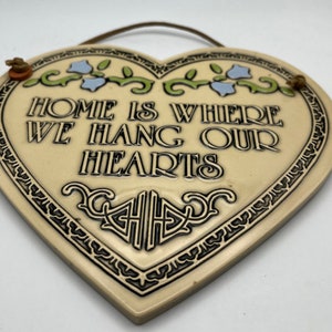 May include: A heart-shaped ceramic plaque with a black and white border and a floral design. The plaque reads "Home is where we hang our hearts".