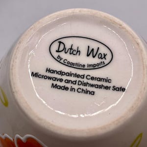 May include: Close-up of the bottom of a ceramic mug. The mug is off-white with colorful floral designs. The base features the text "Dutch Wax by Coastline Imports" and "Handpainted Ceramic Microwave and Dishwasher Safe Made in China."