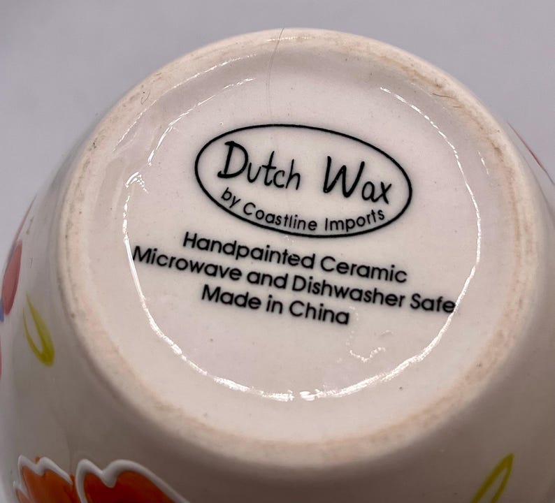 May include: Close-up of the bottom of a ceramic mug. The mug is off-white with colorful floral designs. The base features the text "Dutch Wax by Coastline Imports" and "Handpainted Ceramic Microwave and Dishwasher Safe Made in China."