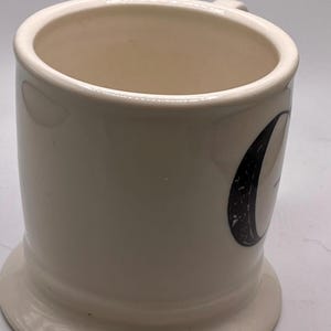 May include: Cream-colored ceramic mug with a rounded base and a small handle. The mug features a black and white graphic on the side. The interior of the mug is a light beige color.
