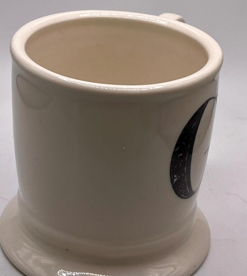 May include: Cream-colored ceramic mug with a rounded base and a small handle. The mug features a black and white graphic on the side. The interior of the mug is a light beige color.