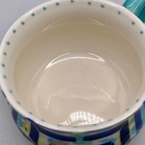 May include: A ceramic mug with a white interior and a turquoise handle. The rim is decorated with small turquoise dots. The exterior features a blue, green, and white geometric pattern. This mug is suitable for hot or cold beverages.
