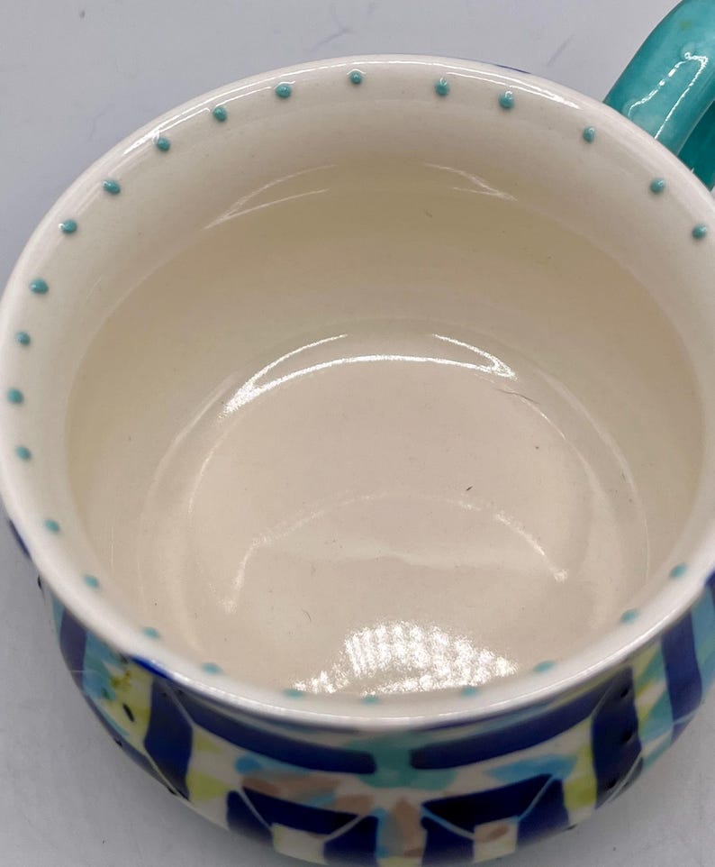 May include: A ceramic mug with a white interior and a turquoise handle. The rim is decorated with small turquoise dots. The exterior features a blue, green, and white geometric pattern. This mug is suitable for hot or cold beverages.