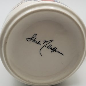 May include: The bottom of a white ceramic mug with a black signature that reads "Karla M. Halfmann".