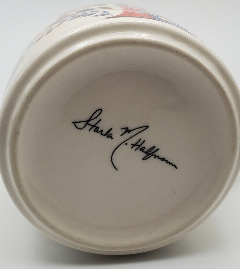 May include: The bottom of a white ceramic mug with a black signature that reads "Karla M. Halfmann".