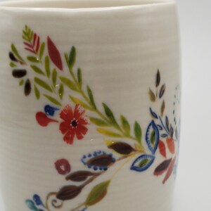 May include: White ceramic mug with a floral design in shades of red, green, blue, and brown. The design features leaves, flowers, and swirls.