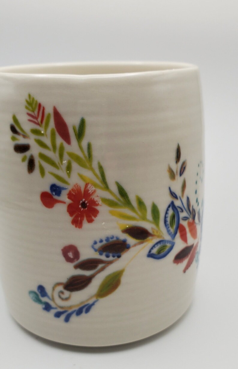 May include: White ceramic mug with a floral design in shades of red, green, blue, and brown. The design features leaves, flowers, and swirls.