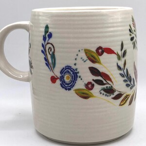 May include: Cream-coloured ceramic mug with a floral design. The design includes blue, red, yellow, and green flowers and leaves. The mug has a textured surface and a curved handle.