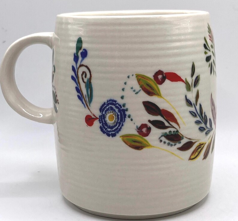 May include: Cream-coloured ceramic mug with a floral design. The design includes blue, red, yellow, and green flowers and leaves. The mug has a textured surface and a curved handle.