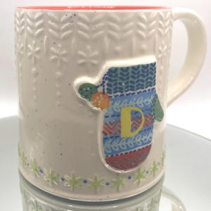 May include: Cream-colored ceramic mug with a red interior and a white handle. The mug features a raised mitten design with the letter "D" and snowflake and leaf patterns. The base has a green and blue snowflake design.
