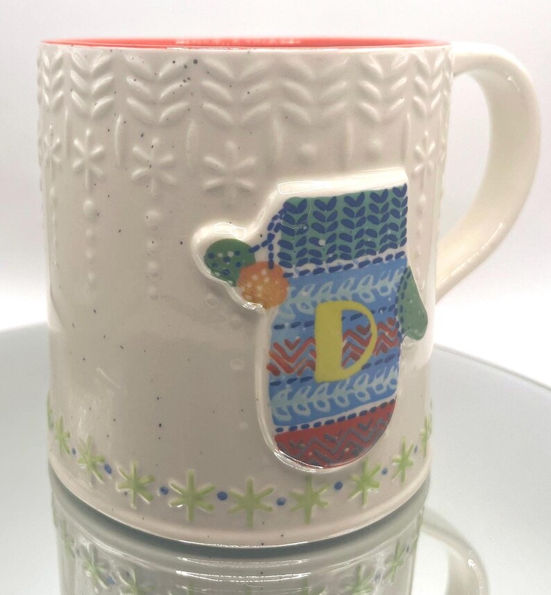 May include: Cream-colored ceramic mug with a red interior and a white handle. The mug features a raised mitten design with the letter "D" and snowflake and leaf patterns. The base has a green and blue snowflake design.