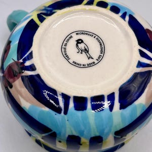 May include: A ceramic mug with a colorful abstract design in blue, yellow, teal, and maroon. The bottom of the mug has a white circle with a bird logo and the text "MICROWAVE & DISHWASHER SAFE MADE IN CHINA".