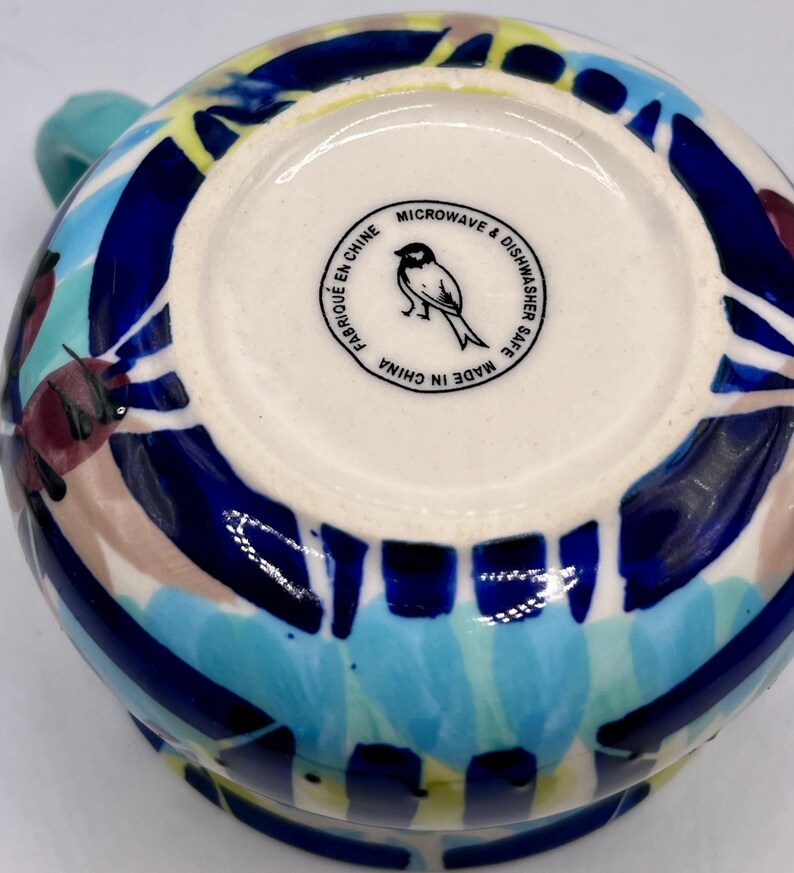 May include: A ceramic mug with a colorful abstract design in blue, yellow, teal, and maroon. The bottom of the mug has a white circle with a bird logo and the text "MICROWAVE & DISHWASHER SAFE MADE IN CHINA".