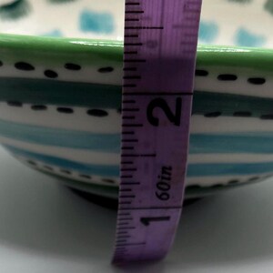 May include: A hand-painted ceramic bowl with a green rim and a design of blue and white stripes. The bowl is approximately 2 inches (5 cm) tall. The interior features a pattern of blue and green squares.