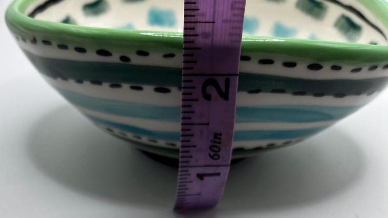 May include: A hand-painted ceramic bowl with a green rim and a design of blue and white stripes. The bowl is approximately 2 inches (5 cm) tall. The interior features a pattern of blue and green squares.