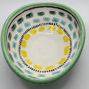 May include: A small, hand-painted ceramic bowl with a white interior. The rim is green, and the exterior features a pattern of blue, green, and yellow shapes. A black dotted line and a black line border the interior design.