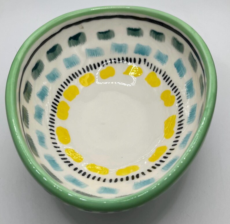 May include: A small, hand-painted ceramic bowl with a white interior. The rim is green, and the exterior features a pattern of blue, green, and yellow shapes. A black dotted line and a black line border the interior design.