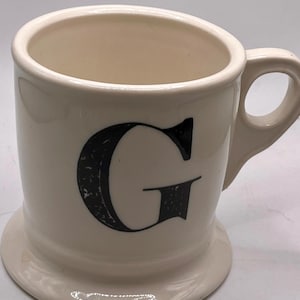 May include: A white ceramic mug with a black handle and the text "But First, Coffee" in a bold, black font. The mug sits on a white surface, showcasing its clean design.