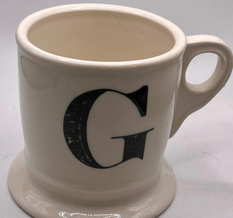 May include: A white ceramic mug with a black handle and the text "But First, Coffee" in a bold, black font. The mug sits on a white surface, showcasing its clean design.