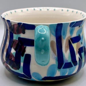 May include: A ceramic mug with a turquoise handle and a white interior. The exterior features a colorful abstract design with blue, navy, maroon, and tan shapes. The rim has small, evenly spaced dots.