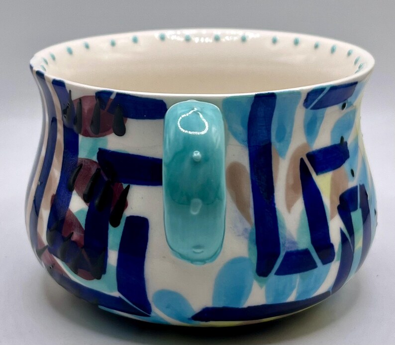 May include: A ceramic mug with a turquoise handle and a white interior. The exterior features a colorful abstract design with blue, navy, maroon, and tan shapes. The rim has small, evenly spaced dots.