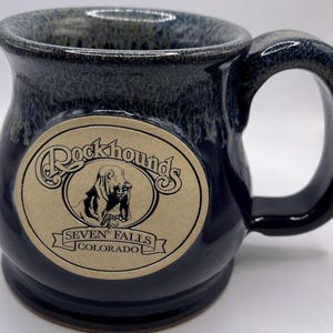 May include: A dark blue ceramic mug with a large handle. The mug features a cream-coloured oval with the words "Rockhounds" and a dog illustration, with "Seven Falls Colorado" below.
