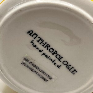 Puede incluir: Un objeto de cer&aacute;mica blanca con el texto "ANTHROPOLOGIE hand painted" en escritura negra. Una etiqueta dice "DISHWASHER & MICROWAVE SAFE MADE IN CHINA / FABRIQUE EN CHINE". El objeto tiene un borde amarillo.