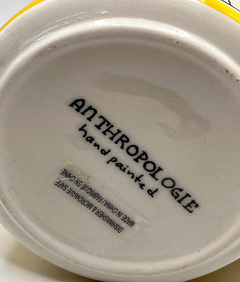 Puede incluir: Un objeto de cer&aacute;mica blanca con el texto "ANTHROPOLOGIE hand painted" en escritura negra. Una etiqueta dice "DISHWASHER & MICROWAVE SAFE MADE IN CHINA / FABRIQUE EN CHINE". El objeto tiene un borde amarillo.