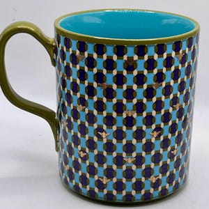 May include: A ceramic mug with a geometric pattern in blue, navy, and olive green, adorned with gold bee accents. The interior is a bright turquoise, and the handle is olive green. A decorative kitchenware item.