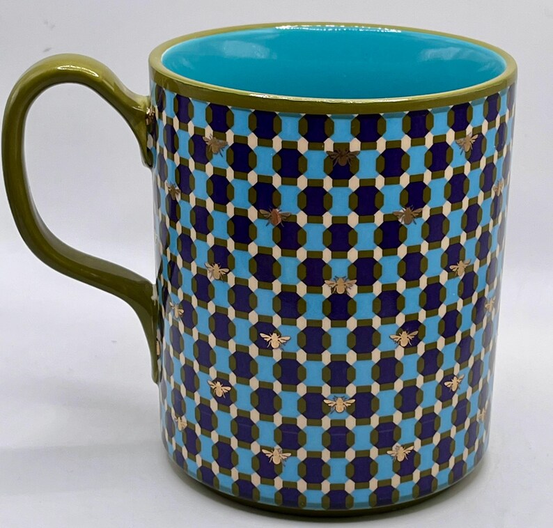 May include: A ceramic mug with a geometric pattern in blue, navy, and olive green, adorned with gold bee accents. The interior is a bright turquoise, and the handle is olive green. A decorative kitchenware item.