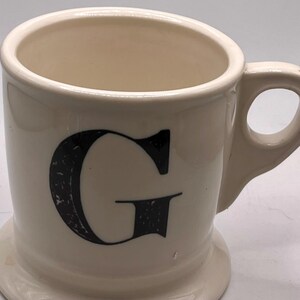 May include: A white ceramic mug with a black handle and the text "But First, Coffee" in a black, handwritten-style font. The mug is on a white surface.