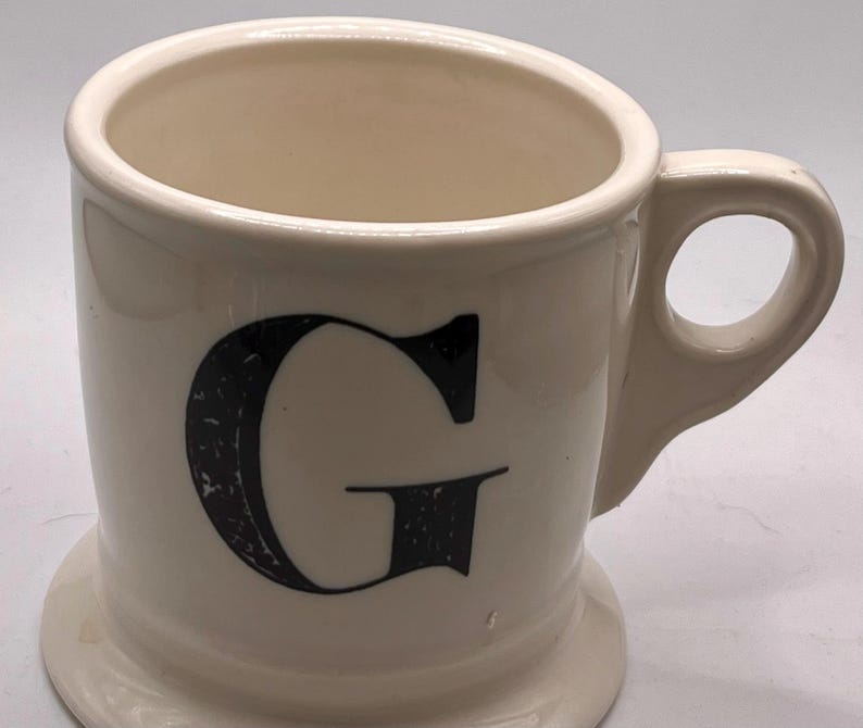 May include: A white ceramic mug with a black handle and the text "But First, Coffee" in a black, handwritten-style font. The mug is on a white surface.