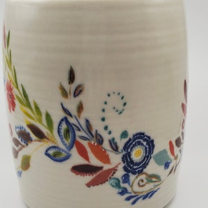 May include: White ceramic mug with a floral design in shades of blue, green, red, and yellow. The design features stylized flowers and leaves.