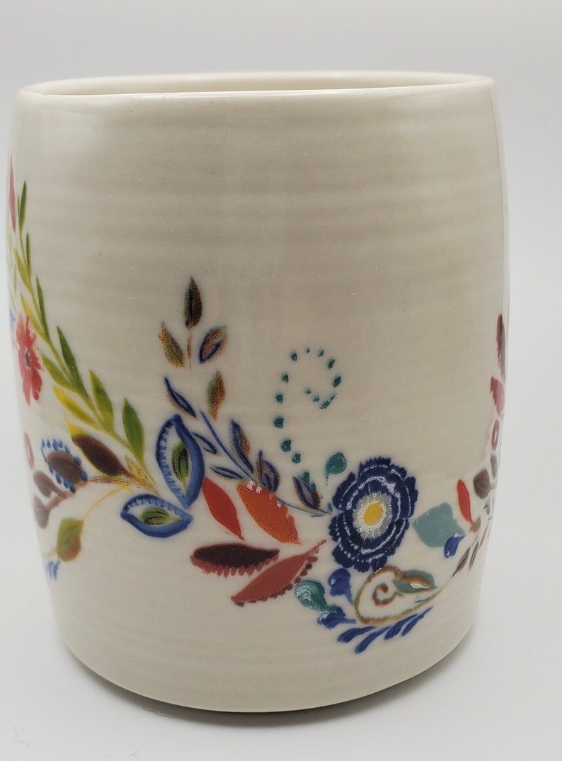 May include: White ceramic mug with a floral design in shades of blue, green, red, and yellow. The design features stylized flowers and leaves.