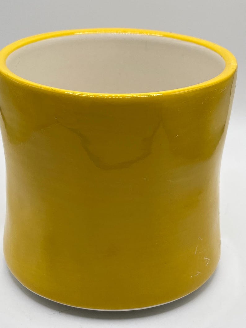 Puede incluir: Una taza de cer&aacute;mica amarilla con acabado brillante. La taza tiene forma cil&iacute;ndrica con un borde ligeramente ensanchado y un interior color crema. El exterior es de color amarillo brillante.
