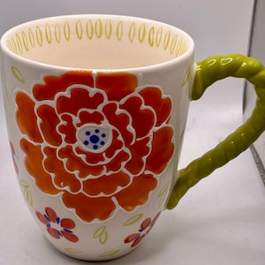 May include: Cream-colored ceramic mug with a large orange and red floral design. The mug features a green handle and smaller red and blue floral accents. The rim is decorated with small yellow dashes. A decorative and colorful coffee mug.