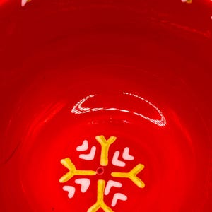 Puede incluir: Un cuenco de cer&aacute;mica rojo brillante con un dise&ntilde;o de copo de nieve en el centro. El copo de nieve es amarillo y blanco. El borde del cuenco tiene una serie de detalles blancos y amarillos. Ideal para servir comida o como pieza decorativa.