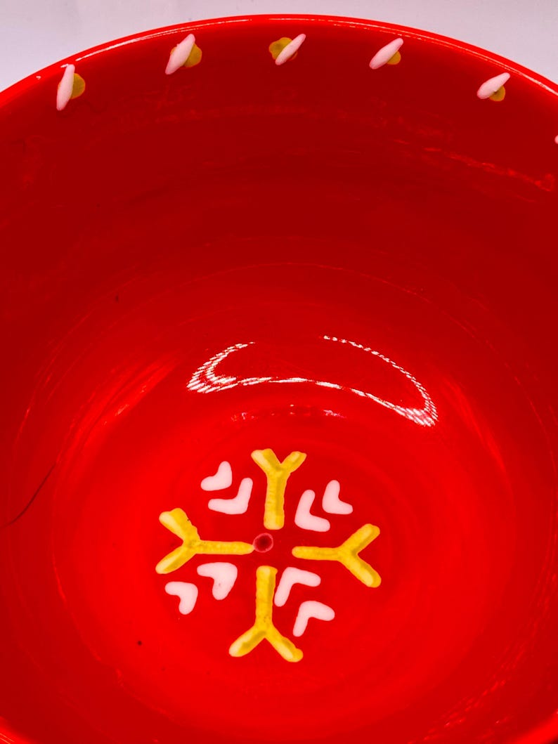 Puede incluir: Un cuenco de cer&aacute;mica rojo brillante con un dise&ntilde;o de copo de nieve en el centro. El copo de nieve es amarillo y blanco. El borde del cuenco tiene una serie de detalles blancos y amarillos. Ideal para servir comida o como pieza decorativa.