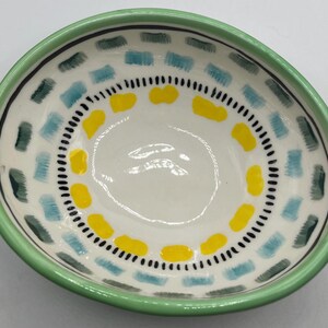 May include: A small, hand-painted ceramic bowl with a white interior. The bowl features a green rim and is decorated with blue, yellow, and green rectangular shapes and black dots. The bowl is ideal for serving snacks or small portions.