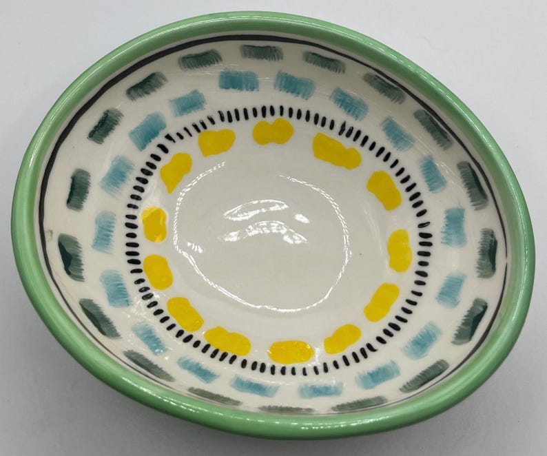 May include: A small, hand-painted ceramic bowl with a white interior. The bowl features a green rim and is decorated with blue, yellow, and green rectangular shapes and black dots. The bowl is ideal for serving snacks or small portions.