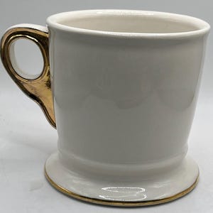 May include: White ceramic shaving mug with a gold-trimmed handle and base. The mug has a wide, round opening and a slightly flared base.